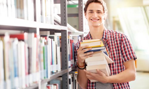 Portrait of a young student holding books standing by bookshelf in library